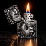 An ornate silver metal lighter with a raised snake design on the front and back, shown open with a flame lit against a dark background. — view 2